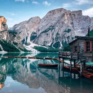 A wooden boathouse on stilts and rowboats on a calm lake, with snow-capped mountains reflected in the water.