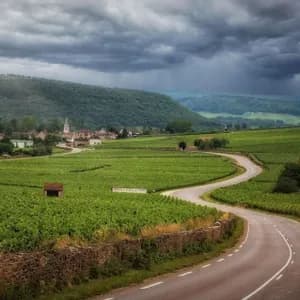 A winding road snakes through lush green vineyards towards a distant village under a sky full of dark storm clouds.