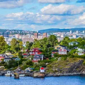 Colorful houses dot a green, rocky shoreline in front of a distant city skyline under a partly cloudy sky.