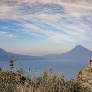 Una persona con un cappello è seduta su una grande roccia, ammirando un vasto lago e montagne lontane sotto un cielo azzurro nuvoloso.