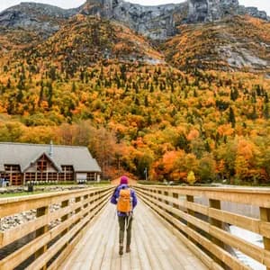 A person with a backpack walks across a wooden bridge towards a large lodge and a mountain covered in colorful autumn foliage.