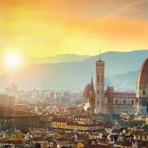 Vista aérea de una ciudad histórica al atardecer, con una gran catedral de cúpula y un alto campanario sobre un fondo montañoso.