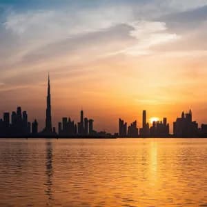 La silhouette di un moderno skyline cittadino contro un vivace tramonto arancione, con i colori del cielo che si riflettono sull'acqua in primo piano.