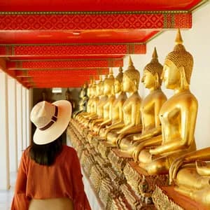A woman wearing a straw hat walks through a temple corridor lined with a long row of golden Buddha statues.