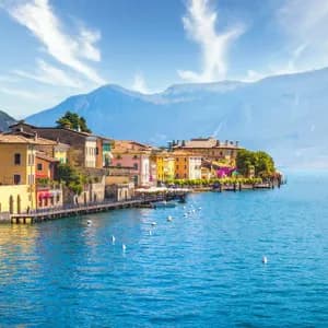 Edifici colorati di una cittadina di lago si specchiano nell'acqua azzurra e brillante, con grandi montagne sullo sfondo sotto un cielo soleggiato.