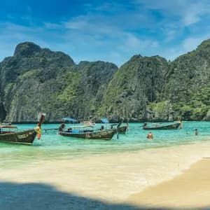 Plusieurs bateaux à longue queue sont amarrés dans une baie turquoise où des gens nagent, avec en arrière-plan de grandes falaises calcaires recouvertes de verdure.
