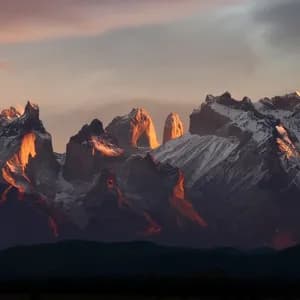 Ein Panoramablick auf eine schneebedeckte Bergkette mit zerklüfteten Gipfeln, beleuchtet vom warmen Licht des Sonnenaufgangs oder Sonnenuntergangs.