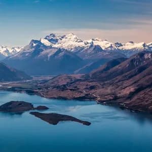 An aerial view of a large blue lake with small islands, surrounded by rugged mountains with snow-capped peaks in the background.