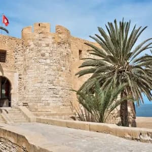 Eine sandfarbene Steinfestung mit einer tunesischen Flagge steht neben einem Strand mit Palmen und dem Meer im Hintergrund.
