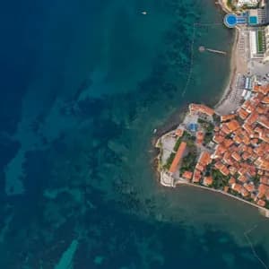 Una veduta aerea, dall'alto, di una città costiera con tetti in tegole arancioni che si affaccia su acque turchesi e limpide, accanto a un porto turistico.