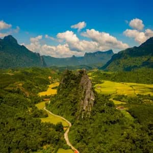 Une vue aérienne d'une route sinueuse à travers une vallée verdoyante avec des rizières, entourée de majestueuses montagnes calcaires sous un ciel bleu.