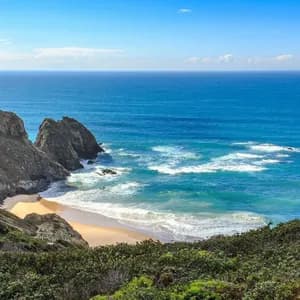 Panorama mozzafiato da una verde scogliera che domina una spiaggia sabbiosa isolata, formazioni rocciose e un vasto oceano blu sotto un cielo limpido.