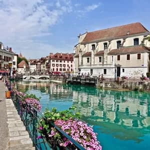 A turquoise canal flows through a historic town, with people strolling on the riverside promenade and buildings reflected in the water.