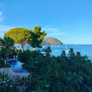 Una piscina immersa nel verde lussureggiante si affaccia su un mare blu con barche e un'isola lontana sotto un cielo sereno.