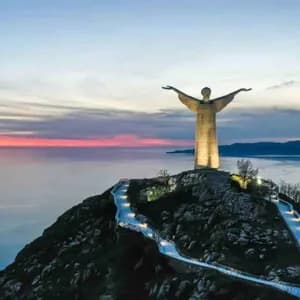 Una grande statua illuminata si erge su una cima di montagna che domina il mare, con un sentiero illuminato che vi conduce al tramonto.