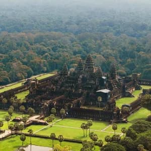 Una vista aerea di un grande e antico complesso di templi in pietra con torri, circondato da prati verdi, palme e una vasta giungla.