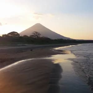 Ein weißer Reiher schreitet an einem feuchten Sandstrand entlang, während Wellen das Ufer sanft umspülen und sich ein großer Vulkan vor dem Sonnenuntergangshimmel abzeichnet.