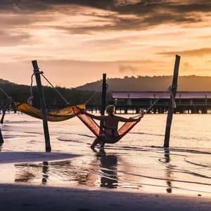 Une personne vue de dos est assise dans un hamac rouge au-dessus de l'eau sur une plage, lors d'un coucher de soleil doré.