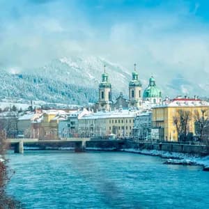 Una vista di una città storica con edifici colorati e cupole di chiese verdi, lungo un fiume, con grandi montagne innevate sullo sfondo.