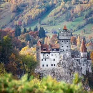 A medieval castle with red-tiled roofs sits on a rocky cliff, nestled in a valley with colorful autumn trees.