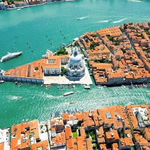Vista aerea di una città con tetti di tegole arancioni e un grande edificio a cupola bianco circondato da acqua turchese e barche.