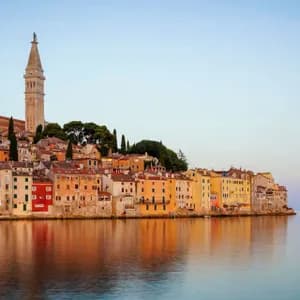 A coastal town with colorful buildings and a prominent bell tower reflecting in the calm water under a clear sky.