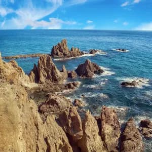 Formaciones rocosas dentadas sobresalen del océano turquesa, vistas desde un acantilado bajo un cielo azul parcialmente nublado.