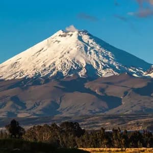 Un gran volcán cubierto de nieve emite una columna de humo desde su cima, dominando un paisaje de colinas y árboles bajo un cielo azul claro.