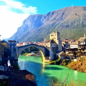 Eine alte Steinbogenbrücke überspannt einen lebhaft türkisfarbenen Fluss, der durch eine historische Stadt fließt, mit einem hohen Berg im Hintergrund.