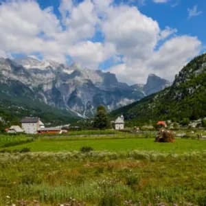 A rural village with a church sits in a lush green valley at the base of jagged, rocky mountains under a partly cloudy sky.