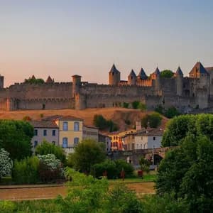 Un grand château de pierre avec de nombreuses tourelles se dresse sur une colline, illuminé par la lumière chaude du coucher de soleil au-dessus d'une petite ville aux arbres verdoyants.