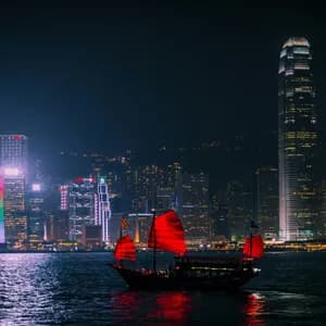 Una tradizionale giunca con vele rosse illuminate galleggia sull'acqua di notte davanti a uno skyline cittadino splendidamente illuminato.