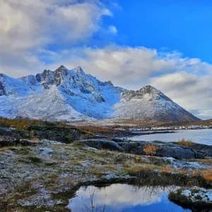 Vista panorámica de montañas nevadas elevándose sobre un pueblo costero y un fiordo, con un primer plano rocoso y parcialmente nevado.