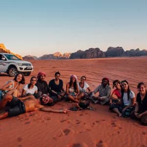 Un grupo de WeRoad se sienta en la arena roja del desierto alrededor de una tetera, con montañas al fondo durante el atardecer.