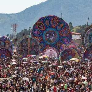 Eine riesige Menschenmenge versammelt sich auf einem Open-Air-Festival, um mehrere riesige, bunte, verzierte Drachen zu bestaunen, die vor einer Bergkette ausgestellt sind.