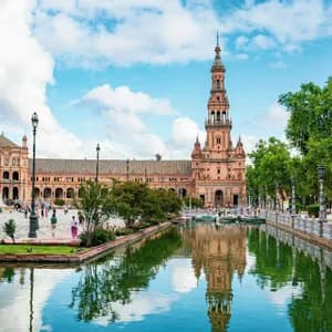 Ein großes, verziertes Gebäude mit einem hohen Turm spiegelt sich in einem Kanal auf einem belebten Platz unter einem bewölkten Himmel.