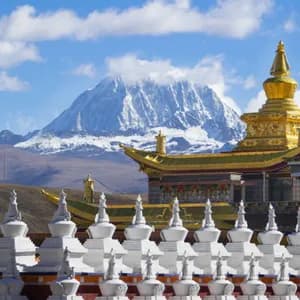 A row of white stupas in front of a golden-roofed temple with a large, snow-covered mountain in the background.