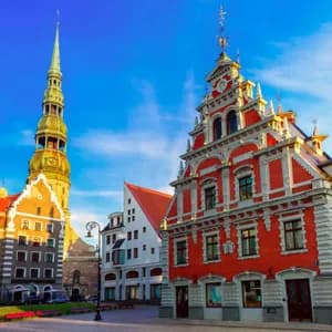 Des bâtiments ornés en briques rouges et un clocher d'église se dressent sur une place de ville sous un ciel bleu clair.
