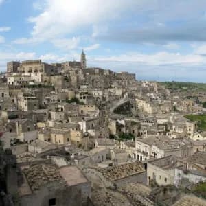 Una vista dall'alto di una città storica con edifici in pietra e tetti in terracotta, costruita sul fianco di una collina sotto un cielo parzialmente nuvoloso.