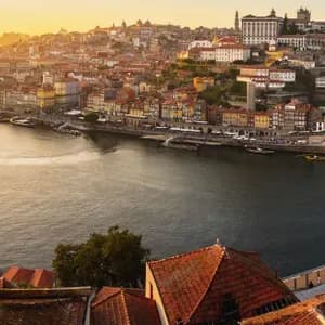 Vue panoramique d'une ville au bord d'une rivière au coucher du soleil, avec un grand pont arqué et des bâtiments colorés à flanc de colline.