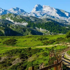 Un sendero de piedra con una valla de madera serpentea a través de un valle de montaña verde con picos rocosos al fondo bajo un cielo despejado.