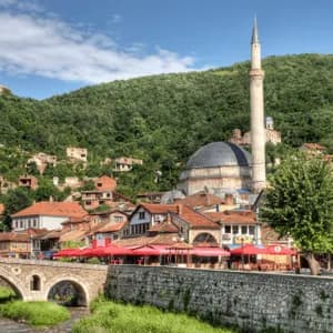 Un ponte di pietra attraversa un fiume in una città storica che ospita una moschea, il tutto incastonato ai piedi di una verdeggiante collina boscosa.