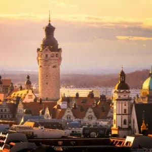 Eine Luftaufnahme einer historischen Stadtkulisse mit einem hohen Steinturm, Kuppeln und Dächern unter einem goldenen Sonnenuntergangshimmel.
