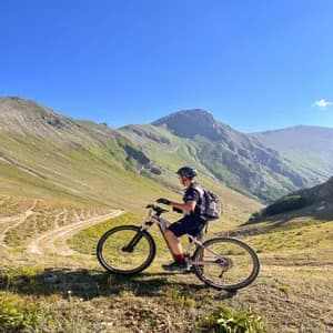 Un mountain biker con il casco si ferma su un pendio erboso, ammirando il panorama di una catena montuosa verde.
