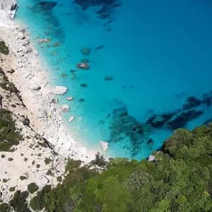 Veduta aerea di una caletta isolata con acqua turchese cristallina, una spiaggia di ciottoli bianchi e scogliere rocciose verdi circostanti.