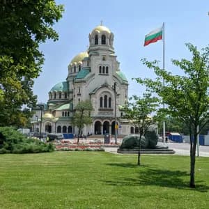 Eine große Kathedrale mit goldenen und grünen Kuppeln und einer bulgarischen Flagge, von einem grünen Park an einem sonnigen Tag aus gesehen.