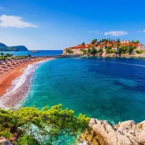 Una vista aerea di un'isola piccola e fortificata con edifici dai tetti rossi, collegata da una strada rialzata a una spiaggia sabbiosa con lettini prendisole.