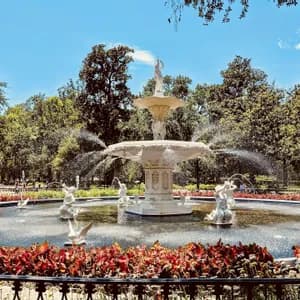 An ornate white tiered fountain with statues spraying water in a public park, with red plants in the foreground and tall trees behind.