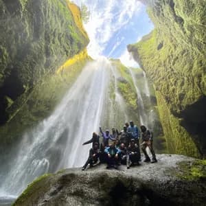 A WeRoad group trip poses on a large rock at the base of a waterfall inside a mossy canyon.