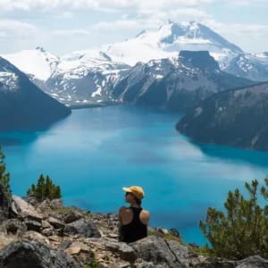 A person in a yellow cap sits on a rocky clifftop, looking out over a bright blue alpine lake surrounded by snow-capped mountains.